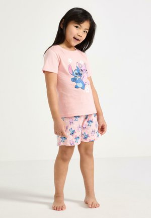 Ung pige i pink T-shirt og shorts, begge med farverige tegneseriefigurer, stående barfodet mod en ensfarvet baggrund.