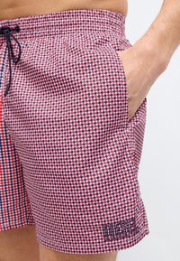 Άνδρας που φοράει σορτς με μοτίβο κουκίδας σε κόκκινο, άσπρο και μπλε χρώμα, με το χέρι στην δεξιά τσέπη. Το λογότυπο Diesel είναι κεντημένο κοντά στο αριστερό τελείωμα.