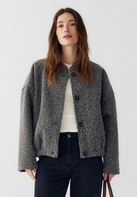 Sivý mäkký vlnený kabát s dlhými rukávmi, okrúhlym výstrihom, štyrmi čiernymi gombíkmi a voľnejším strihom, kombinovaný s bielym topom a tmavými džínsami.