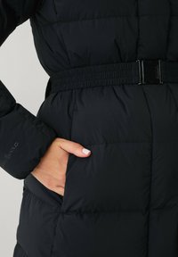 Zwarte pufferjack met een ceintuur in de taille, gestikt textuur, zijzak en elastische manchetten. Het materiaal lijkt glad en geïsoleerd.