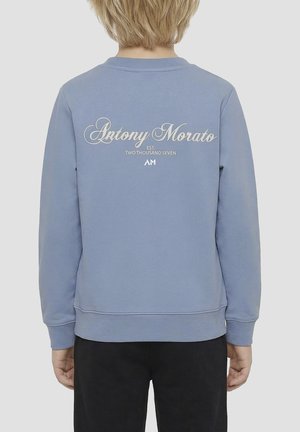 Kind met blond haar draagt een blauwe Antony Morato trui met logo en tekst "EST. TWO THOUSAND SEVEN" op de achterkant, kijkt weg.