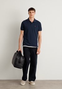 Marine polo skjorte med en liten logo, svarte vide bukser, beige sko og en svart bagasje med håndtak og sidestripper.