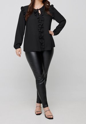 Kobieta ubrana w czarną bluzkę z długim rękawem i falbanami, czarne leginsy ze sztucznej skóry oraz czarne sandały na paski z akcentami z cyrkonii.