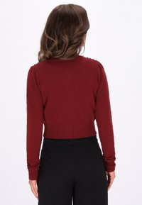 Röd stickad tröja med långa ärmar, ribbad nederkant och dekorativa pärlprydnader vid ärmsluten. Åtsittande, kort design med rund nacke.