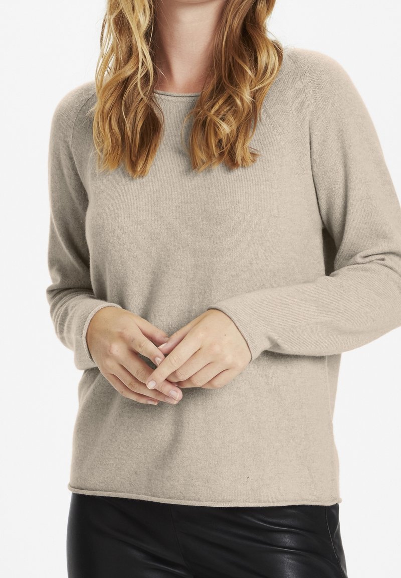 Femme aux cheveux blonds ondulés portant un pull beige à manches longues et un pantalon en cuir noir, les mains doucement jointes devant.