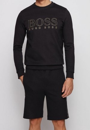 Homme portant un sweat-shirt noir à manches longues Hugo Boss et un short noir assorti, les mains dans les poches.