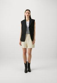 Zwarte mouwloze blazer, witte aansluitende top, crèmekleurige textuurschoenen met bloemenpatroon, kniehoge zwarte leren laarzen, neutrale achtergrond.