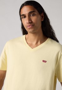 Tricou din bumbac galben cu decolteu în V, cu un mic patch cu logo-ul roșu Levi's pe piept. Textură netedă și mâneci scurte.