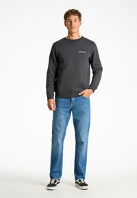 Sudadera gris oscuro con cuello redondo y puños de canalé, combinada con jeans de mezclilla azul claro y zapatillas negras, de pie frente a un fondo blanco.