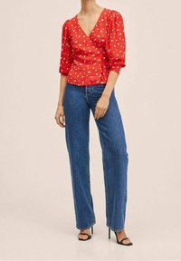 Blouse rouge à manches bouffantes avec un motif floral blanc, décolleté en V profond, associée à un jean droit taille haute bleu et des talons noirs.