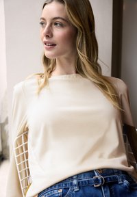 Kremowy, długorękawowy top z prążkowanym dekoltem, zestawiony z dżinsami. Gładka faktura i luźny krój, na tle jasnego tła.