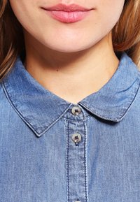 Camicia di denim azzurro chiaro con colletto classico, chiusura a bottoni e un tessuto liscio e testurizzato con dettagli di cucitura sottili.
