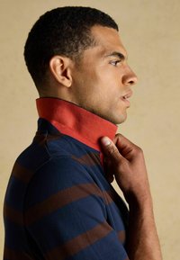 Kreklu ar apkakli, kas ir ar tumši ziliem un brūniem svītrainiem, un kontrastējošu sarkanu apkakli. Audums izskatās teksturēts un elpojo�šs.