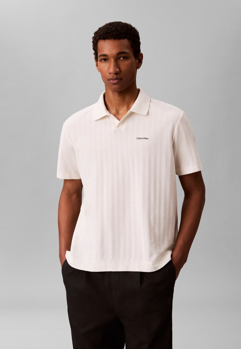 Homme portant un polo blanc manches courtes avec un petit logo sur la poitrine et un pantalon noir, debout les mains dans les poches devant un fond gris.