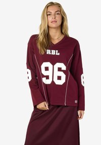 Burgundijska dugosleeve majica s bijelim detaljima i brojevima "96" i "86." Ima V-izrez i opuštenu siluetu, izrađena je od mekane tkanine.