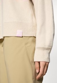 Beige Strickpullover mit geripptem Saum und Bündchen. Mit einem kleinen rosa Etikett am unteren Rand. Kombiniert mit hellbraunen Hosen.