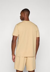 Beige cotton crew-neck t-shirt with short sleeves, worn with matching shorts, viewed from the back against a plain background.