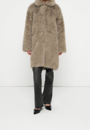 Long manteau en fausse fourrure taupe avec col, porté sur un pantalon noir en cuir à jambes larges et des chaussures noires à bouts pointus avec des nœuds.