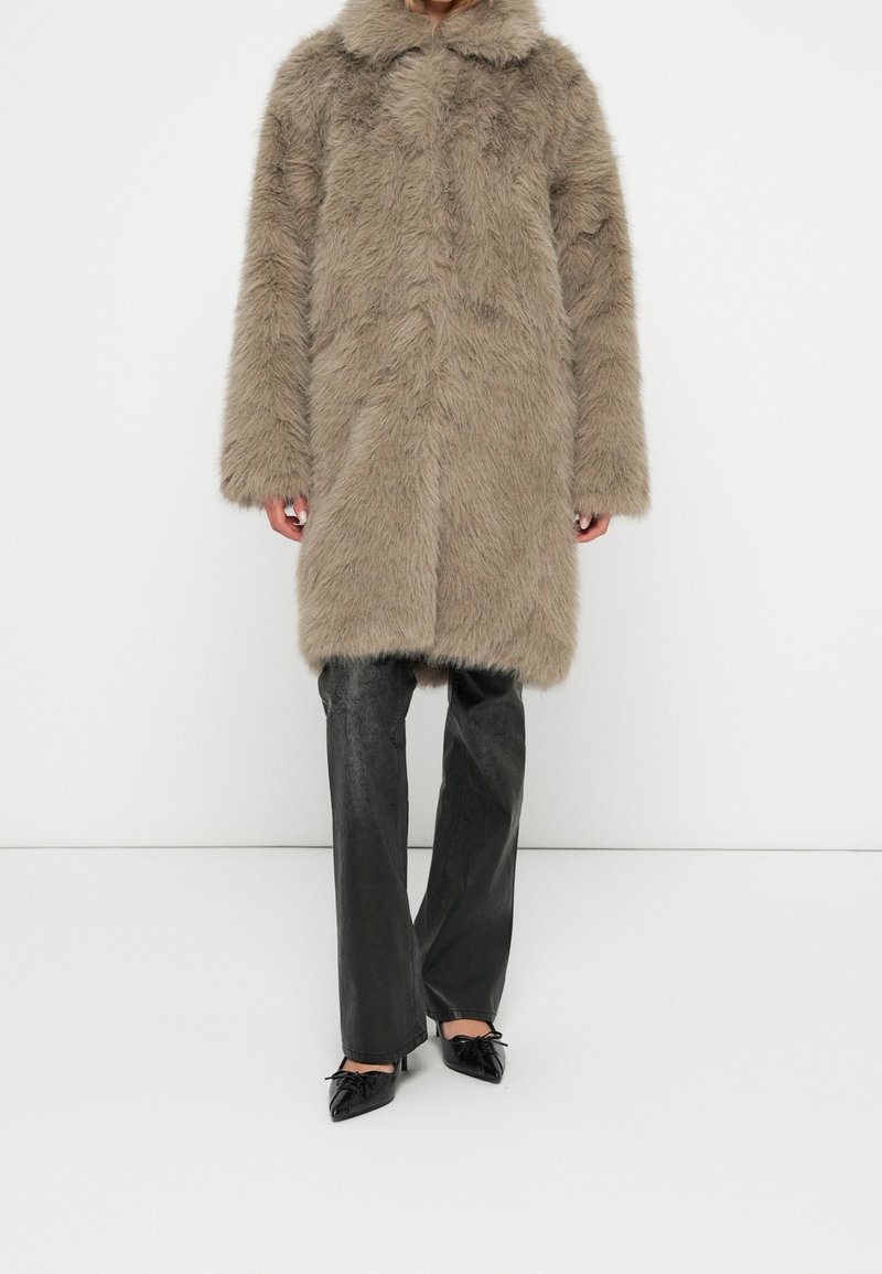 Long manteau en fausse fourrure taupe avec col, porté sur un pantalon noir en cuir à jambes larges et des chaussures noires à bouts pointus avec des nœuds.