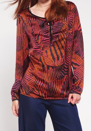 Femme portant un chemisier à manches longues avec des rayures abstraites de zèbre rouges, orange et noires, associé à un pantalon bleu foncé, main reposant près de la hanche.