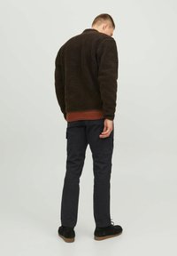 Hnedá fleece bunda s textúrovaným povrchom, červený rebrovaný lem, v kombinácii s tmavými cargo nohavicami. Model stojí obrátený, pričom ukazuje zadný pohľad.