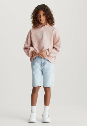Sudadera rosa con un logo, corte holgado, puños de canalé; pantalones cortos de mezclilla azul claro; zapatillas blancas con detalles oscuros; calcetas visibles.