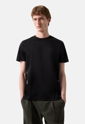 Young man with short brown hair wearing a plain black short-sleeve t-shirt and dark green pants, hands in pockets, neutral background.