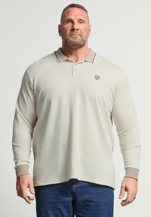 Mann mit getrimmtem Bart, der ein beigefarbenes langärmliges Poloshirt und blaue Jeans trägt, steht vor einem einfarbigen hellen Hintergrund und blickt nach vorne.