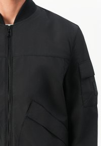 Black bomber jacket featuring a front zipper, ribbed collar, and two side pockets. Includes a patch on the sleeve and a smooth fabric texture.