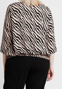 Donna che indossa una blusa ampia a strisce zebrate beige e nere con maniche a tre quarti e pantaloni neri, vista da dietro.