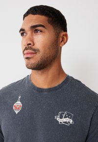 Dark gray long-sleeve shirt with graphic designs: a colorful ornament and playing cards with "M E R R Y X M A S" in white on the chest area.