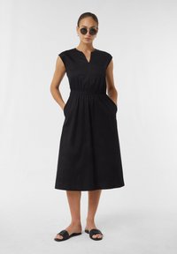 Czarna bawełniana sukienka z krótkimi rękawami, dekoltem w kształcie litery V, elastycznym pasem i bocznymi kieszeniami. Noszona z czarnymi klapkami. Gładka tekstura.