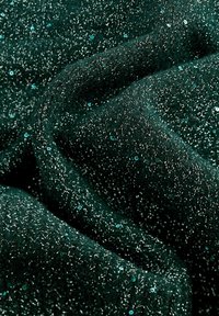 Tissu vert foncé avec des fils argentés brillants et des petites paillettes bleu sarcelle dispersées, montrant des plis et une surface texturée.