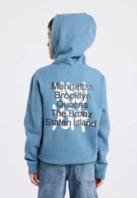 Sweat à capuche bleu clair, avec un grand design de texte graphique gris et blanc au dos. Fabriqué en tissu doux et texturé.