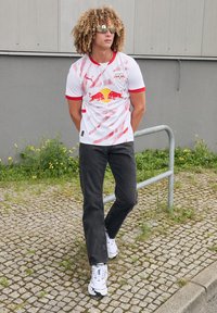 Balta sportinė marškinė su raudonomis akcentais, su įstrižomis juostomis ir geltona/raudona logo. Juodi džinsai ir balti sportiniai bateliai užbaigia įvaizdį.