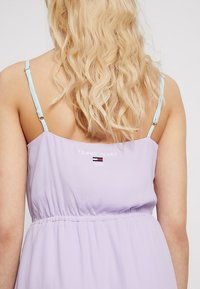 Lilas mekko, jossa on säädettävät vaaleansiniset olkaimet, kootut vyötärö ja brodeerattu "TOMMY JEANS 1985" -logo selässä.