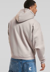 Beige hettegenser med avslappet passform, med ribbestrikkede mansjetter og kant. En logodetalj er synlig på ryggen, sammen med blå denimshorts.