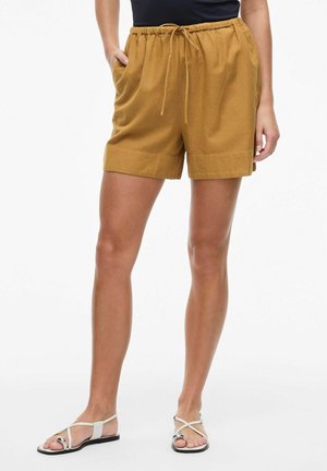 Persona con pantalones cortos mostaza con cordón y bolsillos, y sandalias planas blancas de tiras, de pie contra un fondo liso.