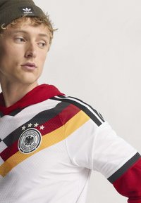 Jeune homme portant un maillot de football allemand avec des rayures noires, rouges et jaunes et un bonnet Adidas vert olive, regardant sur le côté.