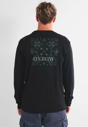 Hombre con pelo corto que lleva una camisa negra de manga larga con un gráfico simétrico verde y el texto "OXBOW" en la parte posterior, combinada con pantalones cargo azules.