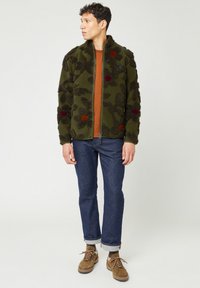 Grön fleecejacka med blomsterstrukturdetaljer, dragkedjestängning och framfickor. Bärs över en orange skjorta, i kombination med blå jeans och bruna stövlar.