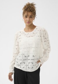 Vrouw met krullend haar, die gouden oorbellen in de vorm van een ring draagt, een witte bloeiende kanten blouse met lange mouwen en zwarte broek aan heeft, staand met een hand in haar zak.