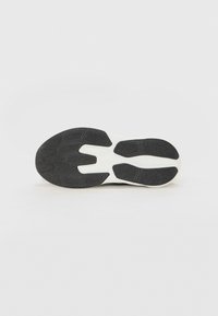 Semelle extérieure en caoutchouc noire avec un motif de grip texturé et une semelle intermédiaire blanche. Présente une forme épurée avec des rainures et des découpes distinctes.