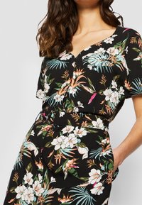 Mujer con un vestido negro de estampado floral, de mangas cortas, escote en V, cinturilla ceñida con un cinturón y una mano en el bolsillo.