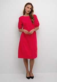 Rochie din bumbac fucsia, cu mâneci bufante, guler rectangular și siluetă până la genunchi; purtată cu tocuri negre; textură netedă, culoare solidă.
