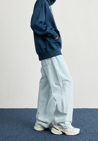 Persona vestida con una sudadera azul marino, jeans de tiro recto de color azul claro y zapatillas blancas con detalles en azul marino y beige, de pie sobre una alfombra azul.