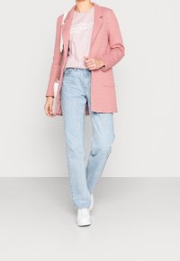 Blazer lungo rosa sopra una t-shirt rosa chiaro con testo, abbinato a jeans blu chiaro e sneakers bianche. Texture lisce e vestibilità sartoriale.
