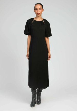 Femme portant une robe midi noire avec un décolleté froncé et des manches courtes élastiquées, associée à des bottes en cuir noires montant jusqu'au genou.