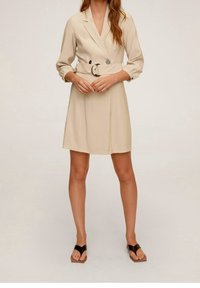 Beige, doppelt geknöpftes Kleid mit langen Ärmeln und tailliertem Gürtel, mit glatter Textur und kniehohem Rock. Schwarze Sandalen.