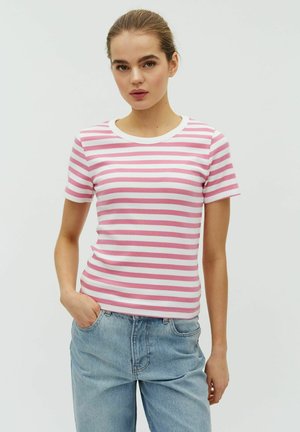 Fiatal nő rózsaszín-fehér csíkos rövid ujjú ingben és világoskék farmerral, egyik kezét a zsebében tartva áll.